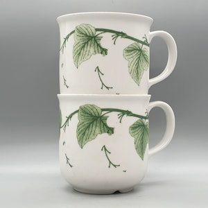 Puede incluir: Dos tazas de cerámica blanca con un diseño de hojas de hiedra verdes. Las tazas están apiladas una encima de la otra.