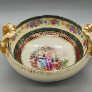 May include: A cream-colored porcelain bowl with two ornate gold handles. The interior features a central scene of two figures and a child, surrounded by a deep red and green border with gold filigree and a rim decorated with colorful floral garlands.