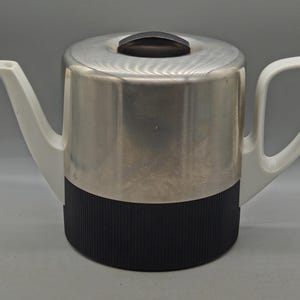 Pode incluir: Uma chaleira de aço inoxidável com uma base preta e canelada e uma alça e um bico brancos. A tampa tem uma pequena alça preta. A chaleira tem um design moderno.