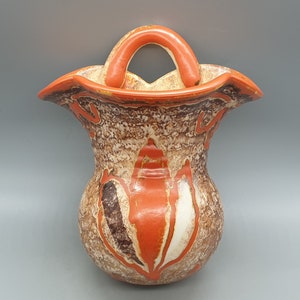 Könnte beinhalten: Eine Vintage-Wandvase mit einer strukturierten, gesprenkelten Oberfläche in Braun-, Creme- und Orangetönen. Die Vase hat einen gebogenen Griff und dekorative orangefarbene Akzente. Das Gesamtdesign erinnert an moderne Keramik aus der Mitte des Jahrhunderts.