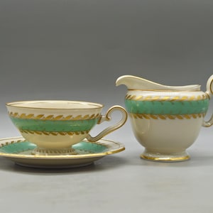 Può includere: Un set di tazza e piattino da tè color crema con una lattiera abbinata. Il set presenta una fascia verde con accenti di foglie d'oro. La tazza e la lattiera hanno finiture dorate. Il piattino è rotondo.