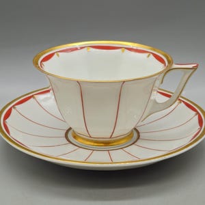 May include: White teacup and saucer set with red and gold accents. The cup and saucer have a gold rim and red vertical lines. The saucer has a gold center and red lines radiating from the center. A classic tea set.