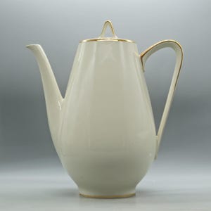 May include: A cream-colored porcelain coffee pot with a gold trim on the rim, handle, and spout. The pot has a tall, slender shape with a lid and a curved handle. The design is simple and elegant.