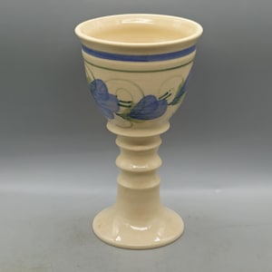May include: A white ceramic goblet with a blue and green floral design. The goblet has a tall, slender stem and a wide, round bowl.