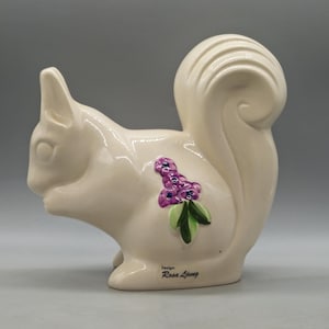 Puede incluir: Figura de ardilla de cerámica color crema con cola rizada. La figura presenta un diseño floral morado y verde en un lateral. La base de la figura tiene el texto "Design: Rosa Ljung".
