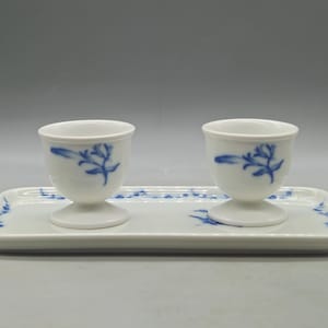May include: Two white porcelain egg cups with blue floral designs, resting on a matching rectangular tray. The tray and cups have a delicate, elegant appearance, suitable for serving eggs. The set is clean and simple.