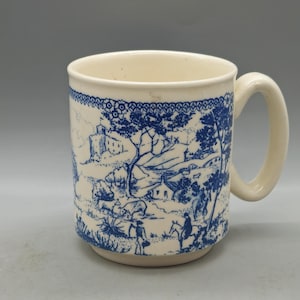Può includere: Una tazza in ceramica bianca con un design blu e bianco di una scena di campagna. La tazza ha un manico e una forma arrotondata.