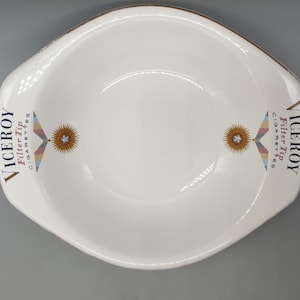 May include: White ceramic ashtray with a gold rim. The ashtray has the words "Viceroy Filter Tip Cigarettes" printed on both sides, along with a stylized logo featuring a star and wings.