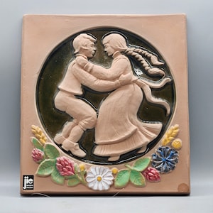Puede incluir: Un azulejo de cerámica con un diseño en relieve de dos personas bailando. El azulejo está decorado con acentos florales verdes, marrones y amarillos. El azulejo está firmado "JIE" en la esquina inferior izquierda.