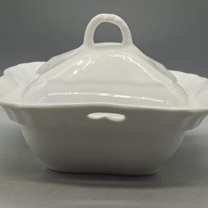 May include: A white ceramic serving dish with a matching lid. The dish features a square shape with scalloped edges and a small cutout handle on the side. The lid has a central arched handle and a smooth, glossy finish throughout the entire piece.