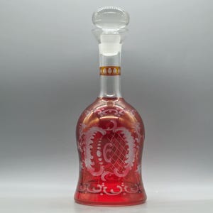 Puede incluir: Una jarra de cristal transparente llena de líquido rojo. Cuenta con un tapón transparente y está adornada con diseños florales blancos grabados. El cuello tiene una banda dorada con un diseño repetido. La jarra es para servir vino o licores.