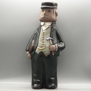 Puede incluir: Una figurita de cerámica de un hombre con una gorra negra y un chaleco verde. Lleva un pico en la mano derecha y tiene un bigote marrón.