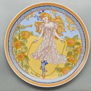 May include: A decorative plate with a design of a girl skipping rope, surrounded by sunflowers. The girl is in a pink dress with white floral details and blue leggings. The plate has a yellow, blue, and orange border, with the artist's signature.