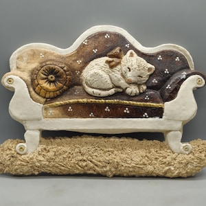 May include: A white ceramic sculpture of a cat sleeping on a brown and white couch with gold accents. The couch is on a tan base.