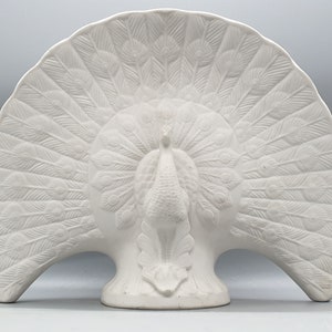May include: White ceramic peacock figurine with a detailed, raised design of feathers. The peacock's tail is spread out in a fan shape.