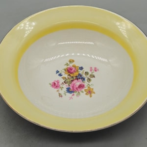 May include: A white ceramic bowl with a yellow rim and a floral design. The bowl has a white background with a yellow rim and a floral design in the centre. The flowers are pink, yellow, and blue.