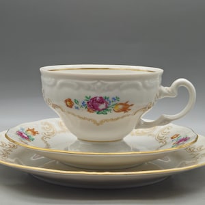 Puede incluir: Juego de taza y platillo de porcelana blanca con diseño floral. La taza y los platos tienen ribetes dorados y un patrón decorativo. El diseño floral incluye rosas rosas, flores azules y detalles en naranja. El juego se muestra sobre un fondo gris.