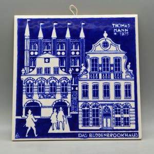 May include: A square, blue and white ceramic tile featuring a detailed illustration of buildings and figures. The tile has the text "THOMAS MANN 1875" and "DAS BUDDENBROOKHAUS". A small loop is attached for hanging.