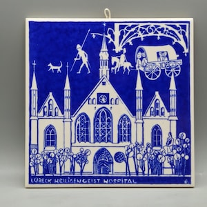 May include: A square, blue and white ceramic tile featuring a detailed illustration of a building with spires, arched windows, and a crowd of people. The tile includes the text "LURECK HEILIGENGEIST HOSPITAL".