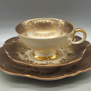 May include: A cream-colored porcelain teacup, saucer, and plate set featuring a metallic gold floral pattern. The teacup has a gold-trimmed handle and base, while the saucer and plate have scalloped edges with a deep brown and gold finish.