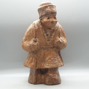 May include: A brown ceramic figurine of a person wearing a hat and a long coat. The figurine is standing with its arms crossed and has a textured surface.