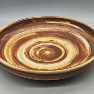 May include: A handmade ceramic bowl with a spiral design. The bowl features concentric circles of brown, tan, and cream colours. The rim and base are a darker brown. This decorative bowl is suitable for serving or display.