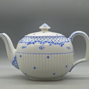 Puede incluir: Tetera de porcelana blanca con diseño floral y enrejado azul. La tetera tiene un pico curvo, un cuerpo redondeado con estrías verticales y una tapa a juego con un adorno. El asa es un lazo curvo.