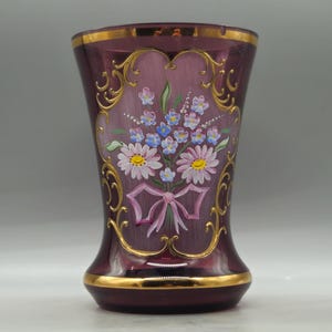 Può includere: Un vaso di vetro bordeaux con finiture dorate e opere d'arte floreali. Il vaso presenta un bouquet dipinto di margherite, nontiscordardimé e un fiocco rosa. Il vaso è alto circa 18 cm.