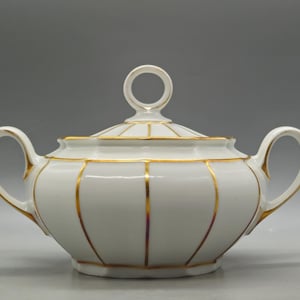 Puede incluir: Azucarero de porcelana blanca con tapa y dos asas. El cuenco está decorado con rayas verticales doradas y ribetes dorados. La tapa tiene un asa de anillo. Diseño clásico.