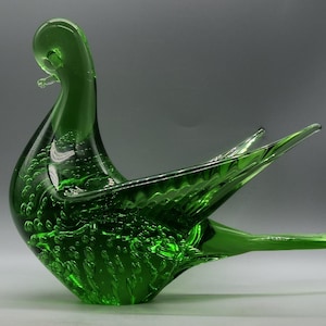 Könnte beinhalten: Eine grüne Glasskulptur einer Taube mit einer strukturierten Oberfläche und eingeschlossenen Luftblasen. Die Taube hat einen langen Hals und eine große Flügelspannweite.
