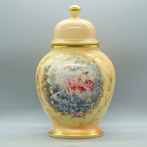 Puede incluir: Un jarrón grande de cerámica de color crema con tapa y ribetes dorados. El jarrón presenta una escena pintada de una mujer en un columpio en un jardín.