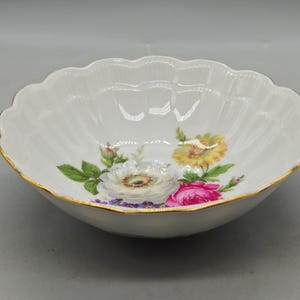 Puede incluir: Cuenco de cerámica blanca con borde festoneado y ribete dorado. El cuenco está decorado con coloridos diseños florales, que incluyen flores blancas, amarillas y rosas con hojas verdes. Un plato decorativo.