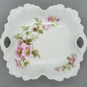 Puede incluir: Un plato de servir de porcelana blanca con dos asas y un diseño floral. El plato presenta rosas rosas y hojas verdes.