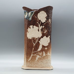 May include: A tall, rectangular ceramic vase with a brown and cream colour scheme. The vase features a floral design and a butterfly accent. The vase is approximately 25 cm tall.