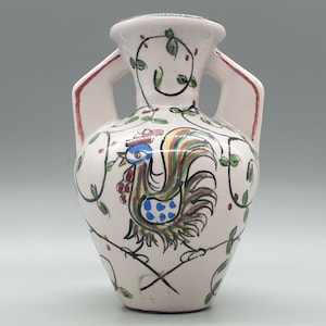 May include: A white ceramic vase with two handles, adorned with a colorful rooster illustration. The vase features red accents on the handles and is decorated with green leaves and vines. The rooster has blue, yellow, and red details.