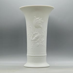May include: A white porcelain vase with a flared top and a detailed floral relief design. The vase features a raised pattern of flowers and leaves, with a decorative band near the base. The vase is a classic piece for displaying flowers.