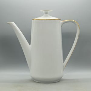 Puede incluir: Una cafetera de porcelana blanca con un pico largo y un asa curvada. La tapa y el asa están adornadas con ribetes dorados. La cafetera tiene una forma alta y esbelta y se asienta sobre una base sólida. La cafetera es probablemente vintage.