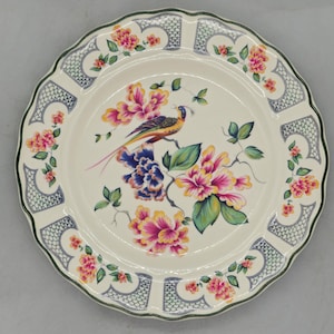 Puede incluir: Un plato decorativo de cerámica con borde festoneado y diseño floral y de pájaros. El plato presenta un pájaro colorido posado en una rama con flores rosas, amarillas y azules. El borde tiene un patrón azul y blanco.