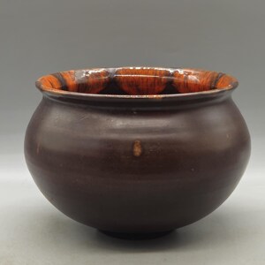 Puede incluir: Un cuenco redondo de cerámica marrón oscuro con acabado brillante. El borde interior presenta un patrón contrastante naranja y marrón. El cuenco tiene una abertura ancha y un cuerpo redondeado, adecuado para uso decorativo o funcional.