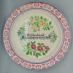 Puede incluir: Un plato de cerámica blanco con un borde rojo y azul. El plato está decorado con un diseño floral y el texto "No weing oder nie, was Gott will ist mein Ziel".