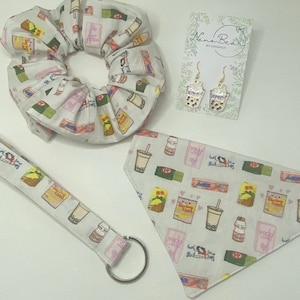 May include: A set of four items featuring a white background with a repeated pattern of bubble tea, snacks, and candy. The set includes a white scrunchie, a white bandana, a white keychain, and a pair of gold earrings with a bubble tea design. The earrings are on a white card with the text "Nana Beads" and "Get Addicted".