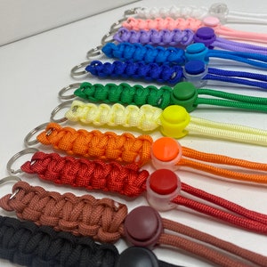 Puede incluir: Una colección de diez llaveros de paracord dispuestos en fila, cada uno con un cordón trenzado de un color distinto, incluyendo blanco, rosa, morado, azul, verde, amarillo, naranja, rojo, marrón y negro, con un tope de plástico a juego.