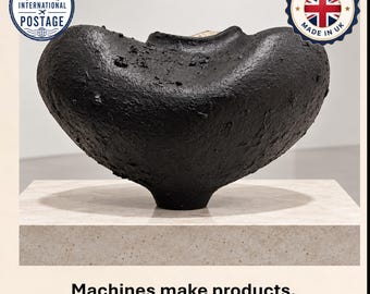 May include: A black, textured, bowl-shaped vessel with a wide, curved rim and a narrow base. The surface has a rough, uneven finish. Below the object, the text reads: Machines make products. Hands create character. The object is centered.
