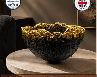 May include: A bowl with a rough, textured black exterior and a metallic gold interior rim. The bowl features an irregular, jagged edge and a deep, rounded basin. The surface of the bowl has a craggy, uneven appearance throughout the exterior.