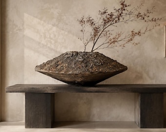 May include: A wide, low-profile bowl with a dark, heavily textured, rugged surface resembling volcanic rock. The bowl has a sharp, angular rim and a tapered base. A delicate, thin, brown branch with small, dried buds extends upward from the center.