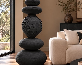 May include: A tall, vertical sculpture composed of five stacked, rounded, charcoal-black stones. The surface of each stone has a rough, textured finish, with the central stone featuring a particularly craggy, uneven appearance.