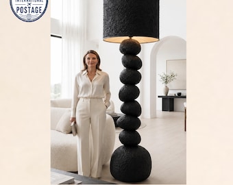 May include: A tall floor lamp featuring a stack of seven rounded, textured black stones forming the base and central column, topped with a large, cylindrical black lampshade with a matching rough, organic surface texture.