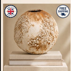 May include: A spherical vase with a textured, off-white and tan surface featuring small, embedded stone-like particles. The vase has a circular opening at the top and rests on a stack of two rectangular, light-coloured books.