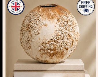 May include: A spherical vase with a textured, off-white and tan surface featuring small, embedded stone-like particles. The vase has a circular opening at the top and rests on a stack of two rectangular, light-colored books.