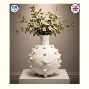 May include: A white spherical vase with a textured, matte surface and raised circular bumps across its body. The vase has a narrow neck and holds a bouquet of white flowers with green leaves. The overall appearance is monochromatic and tactile.